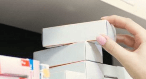 A hand placing a white box on a shelf filled with various medication packages.